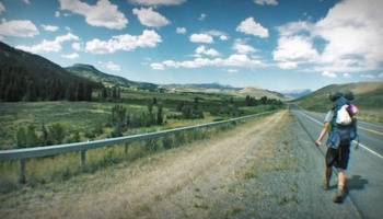 A person carrying a large backpack walks along a rural paved highway through a sunny mountain valley.