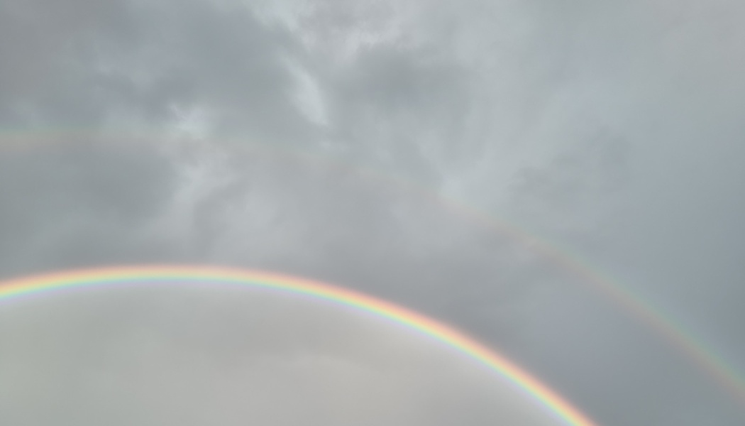 A bright rainbow and a faint secondary rainbow arching across a cloudy, overcast sky.