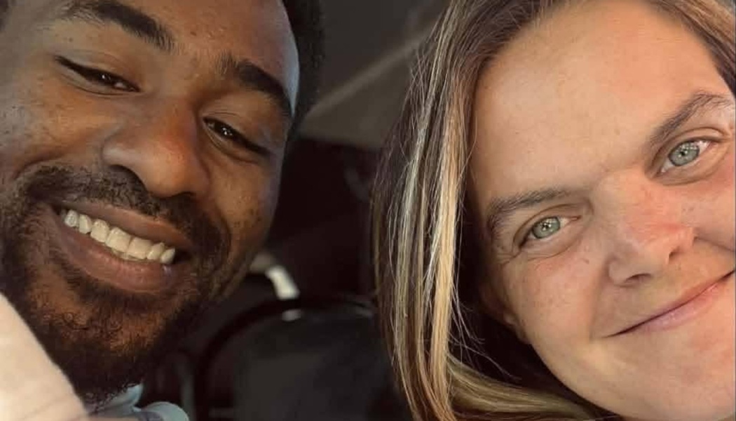 Close-up selfie of a smiling Black man with a beard and a blonde woman smiling next to him.