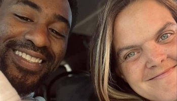 Close-up selfie of a smiling Black man with a beard and a blonde woman smiling next to him.