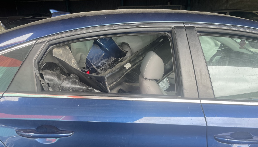 Side view of a blue car with a missing rear window and broken vehicle parts stuffed into the back seat.