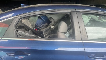 Side view of a blue car with a missing rear window and broken vehicle parts stuffed into the back seat.