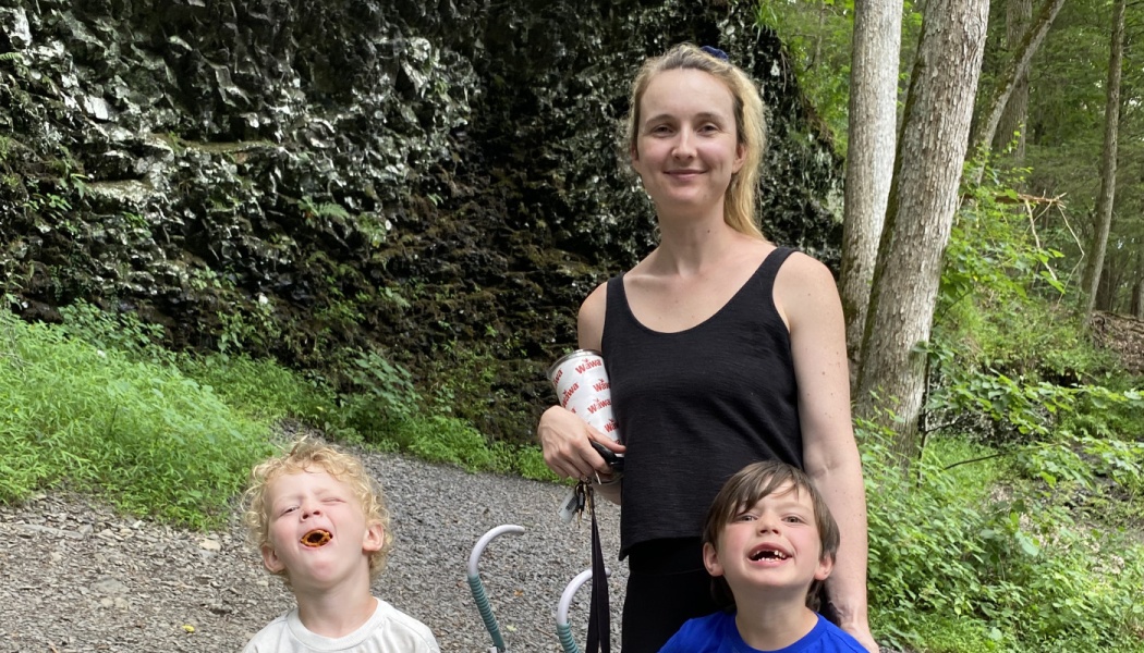 A smiling mother stands outdoors with her two young children making silly faces on a trail by a rocky cliff.