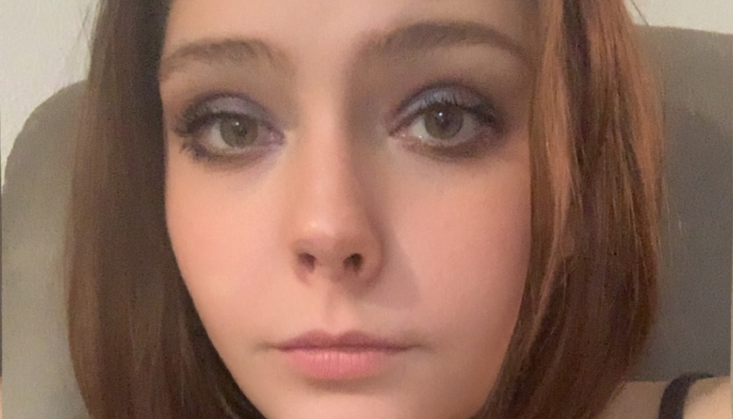 Close-up portrait of a young woman with reddish-brown hair and eye makeup looking directly at the camera.