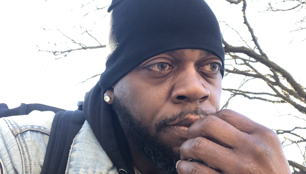 A thoughtful Black man wearing a black beanie and denim jacket, holding his hand near his mouth while outdoors.