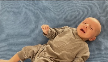 A smiling newborn baby wearing a brown onesie lying on a blue knit blanket.
