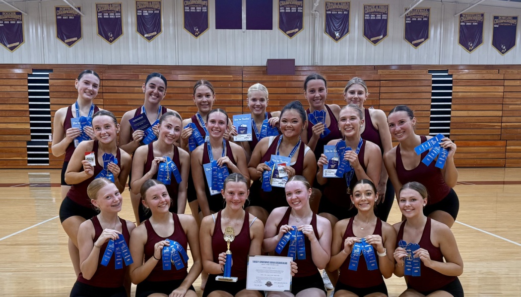 Members of a dance team in maroon uniforms pose with blue award ribbons and a trophy on a basketball court.
