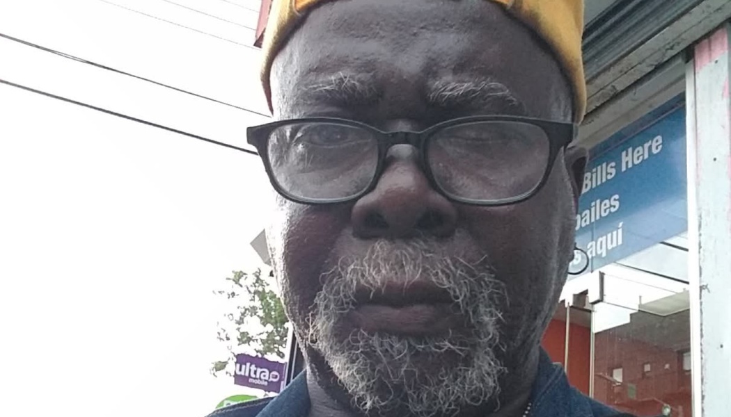 Close-up portrait of an older Black man with black glasses, a grey beard, and a yellow cap outdoors.