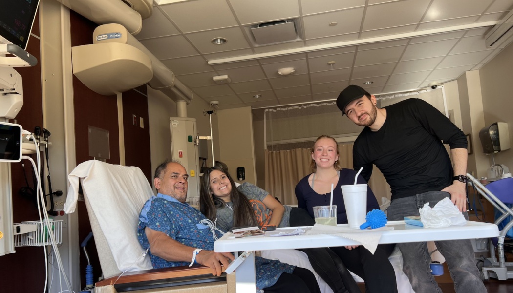 A hospital patient in a blue gown smiles surrounded by three supportive visitors in his hospital room.