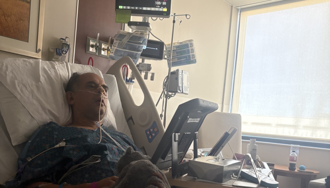 A man with a nasal cannula rests in a hospital bed surrounded by medical monitoring equipment.