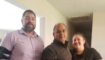 Three smiling adults standing together indoors, including a man wearing a black medical arm brace.