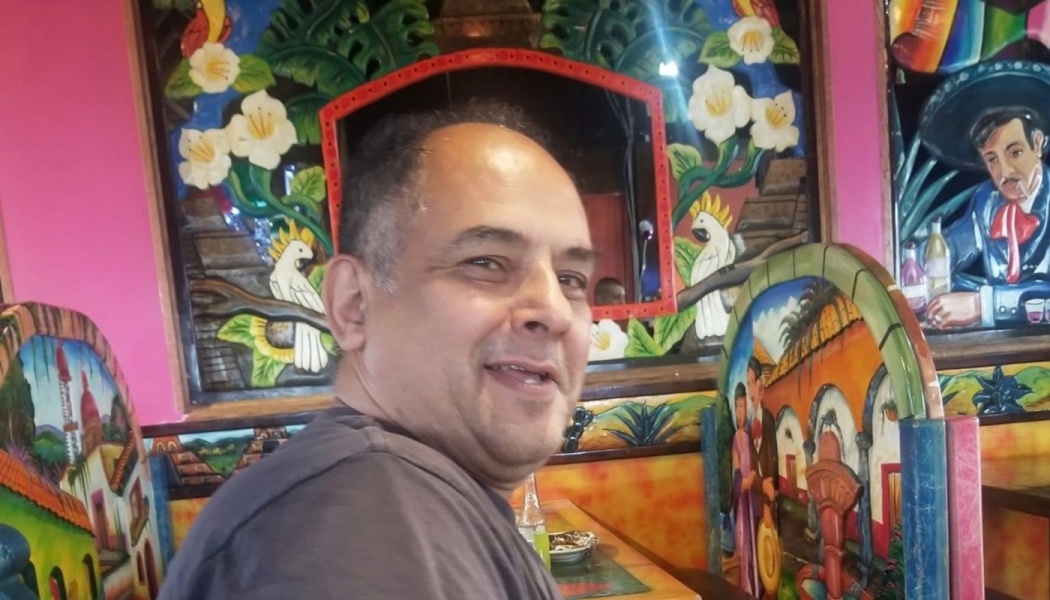 A smiling man sitting in a vibrantly decorated restaurant surrounded by colorful Mexican artwork.