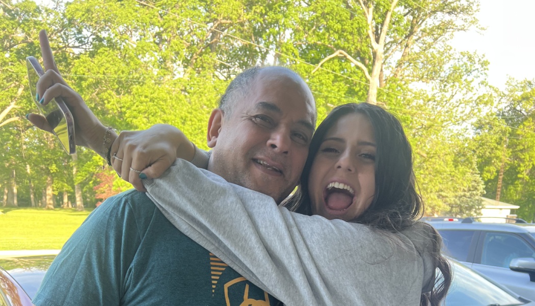 A young woman cheerfully hugs a smiling middle-aged man tightly outdoors with lush green trees in the background.