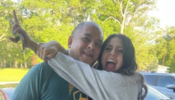 A young woman cheerfully hugs a smiling middle-aged man tightly outdoors with lush green trees in the background.