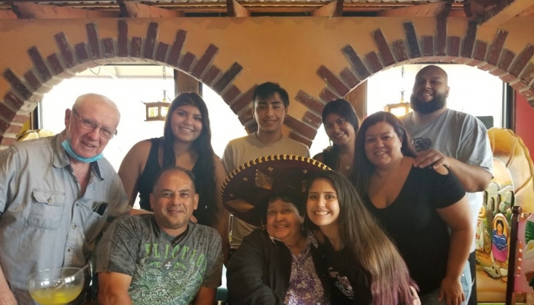A smiling family of nine poses together around an elderly woman wearing a large sombrero at a restaurant.