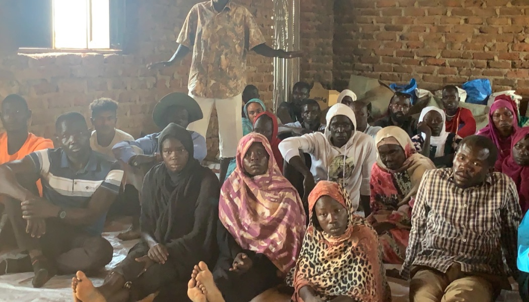 A group of men and women sitting on the floor inside a brick building with a man standing behind them.