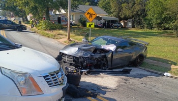 A dark gray sports car with severe front-end damage following a head-on crash with a white car on a street.