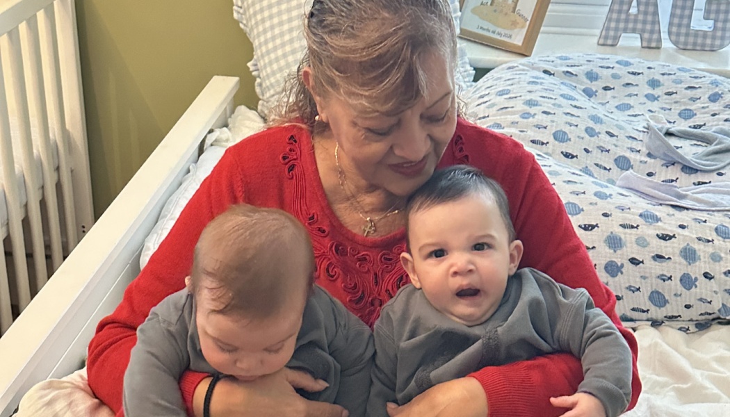 A smiling woman in a red sweater lovingly holds twin babies wearing matching gray shirts.
