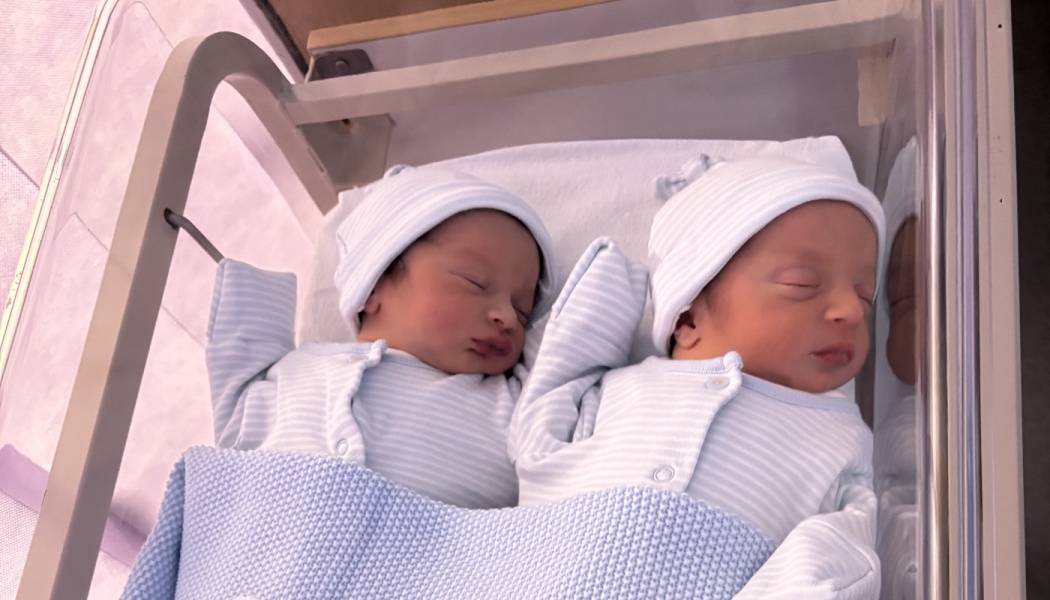 Newborn twin babies sleeping side-by-side in matching light blue striped caps and onesies inside a hospital bassinet.