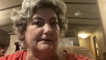 Close-up portrait of a woman with short, wavy grey hair wearing an orange shirt indoors.