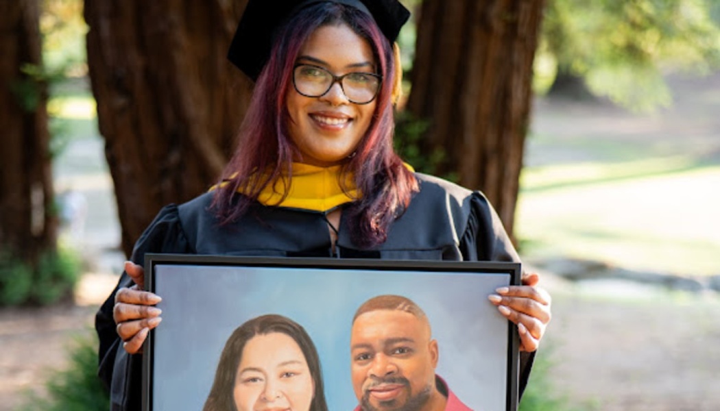 A smiling graduate in a cap and gown holds a framed photo of a man and woman outdoors.