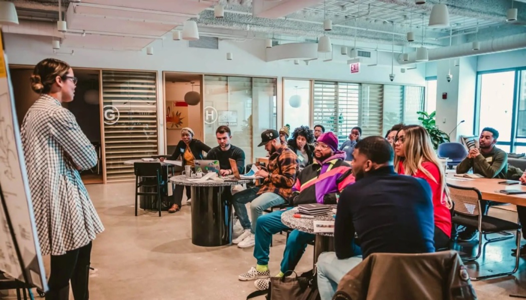 A woman leads a presentation to a diverse audience seated at small tables in a modern office space.