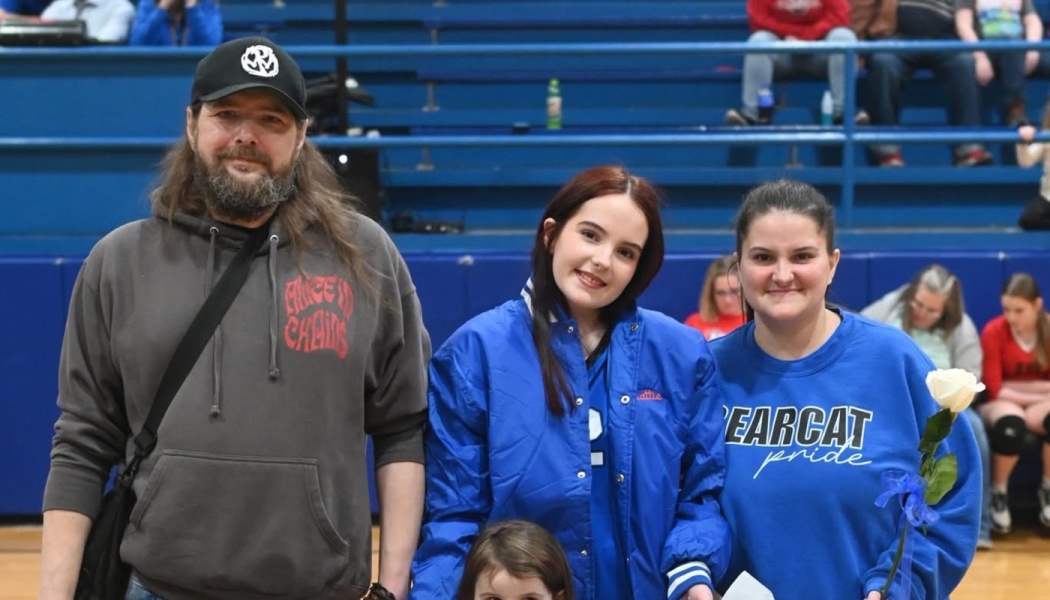 A man, a young woman, and a woman holding a single white rose pose together in a gymnasium in front of blue bleachers.