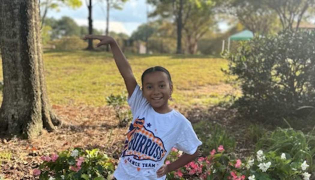 A smiling young girl in a Burruss Dance Team t-shirt striking a dance pose outdoors surrounded by flowers.