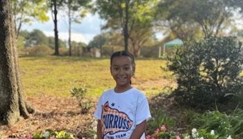 A smiling young girl wearing a white graphic t-shirt standing outdoors in a park with trees.