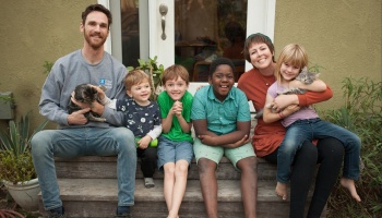 A smiling family of six sitting on wooden porch steps outside, with four children and two cats.
