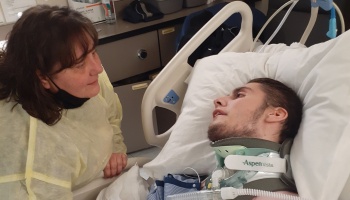 A woman in a medical gown looks tenderly at a young man wearing a neck brace in a hospital bed.