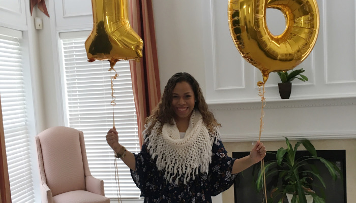 Smiling woman in a fringed white scarf holding large gold foil 18 balloons.