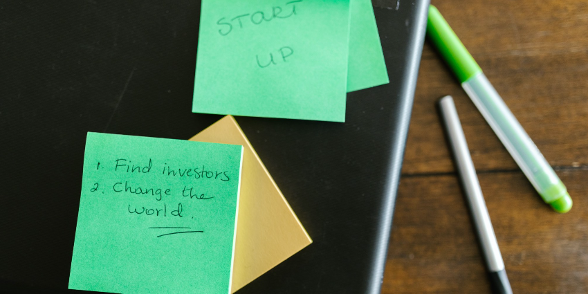 Green sticky notes on a desk that read "Startup" and "1. Find Investors 2. Change the world" to represent 45-110 coming into effect