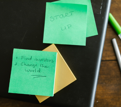 Green sticky notes on a desk that read "Startup" and "1. Find Investors 2. Change the world" to represent 45-110 coming into effect