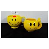Two smiley face mugs, one is showing the peace
