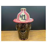 BR2…Steve Turpin Southern folk art face jugs, sculpted with exaggerated features such as prominent bugged eyes, an expressive grin with visible teeth, and a brown-green glaze finish. The figure is t
