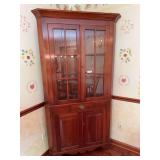 Cherry corner cupboard with upper glass display doors and lower cabinet storage. Features a traditional corner design suitable for dining room, kitchen, or living area display.