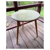 Three-legged table with round glass top and decorative base. Accent table suitable for use on a patio, porch, sunroom, or interior sitting area.