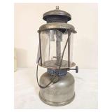 Coleman Quick-Lite pressure lantern marked 10/25, with silver-tone fount and ventilator, clear globe and wire bail handle. Globe shows visible cracking; lantern has age-related oxidation and wear.
