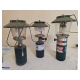 Group of three Coleman LP gas lanterns in assorted styles with green and metal finishes, glass globes and carrying handles; shows varying age-related wear and surface soiling.