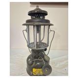 Coleman U.S. Army SMP lantern marked 1963 with dark military-style fount, dark ventilator, clear globe, wire frame and chained cap assembly. Shows substantial paint loss, oxidation and age-related wea