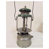 Coleman Model 220 pressure lantern marked 8/28, with silver-tone fount, green porcelain ventilator, clear glass globe and wire bail handle. Shows oxidation and age-related surface wear.