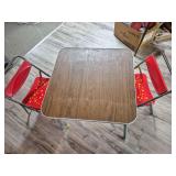 Vintage Child’s Table & Two Metal Chairs. Child-size rectangular table with woodgrain laminate top and metal legs, accompanied by two red metal chairs with yellow splatter decoration.