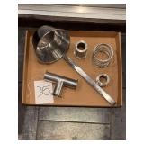 industrial or plumbing hardware, including a ladle-like tool, a T-shaped pipe fitting, a spring, and some small cylindrical connectors.