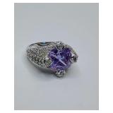 This ring is a statement piece featuring a large, square or cushion-cut purple gemstone. The central stone is set in a wide band, silver-plated metal, and is heavily embellished with smaller clear sto
