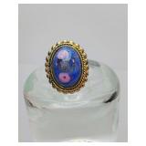 Vintage-style ring featuring an oval-shaped, blue glass stone with pink floral inclusions and a metallic, twisted-rope border setting. The glass has a distinctive, handmade look with small bubbles and