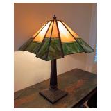 Arts & Crafts style stained glass table lamp, featuring a geometric paneled shade with green and amber tones on a fluted bronze-finish metal base. 22.5"T x 14.5" W at base of shade. Works.