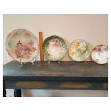 4 cabinet plates left to right: Bavarian Schumann Arzberg "Wild Rose" 11.5". No markings scalloped, molded rim cut-out handles, a deep green, pink roses 10". Bavaria e. Penel yellow roses 8.75". Maker