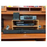 Grouping of electronics - pair of computer speakers, LXI VCR Plus, Msgnavox DVD Recorder, RCA 37” soundbar, Sony DVD Player with remote and wooden stand - none are texted and DVD Recorder has a remo
