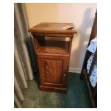 Narrow medium-brown wooden side table featuring an open upper shelf and a paneled cabinet door with a brass-finish knob.